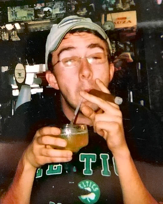 Comedian Joe List of Tuesdays with Stories in a Boston Celtics shirt, with a cigar in one corner of his mouth and a straw to an alcoholic beverage in another. Find more about Joe List at www.tuesdayspod.com/joe-list