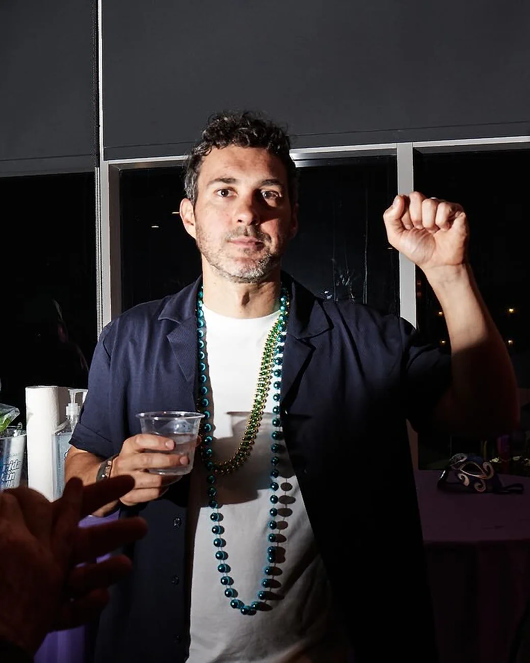 Comedian Mark Normand of Tuesdays with Stories raising his fist while wearing beads. Find more about Mark Normand at www.tuesdayspod.com/mark-normand